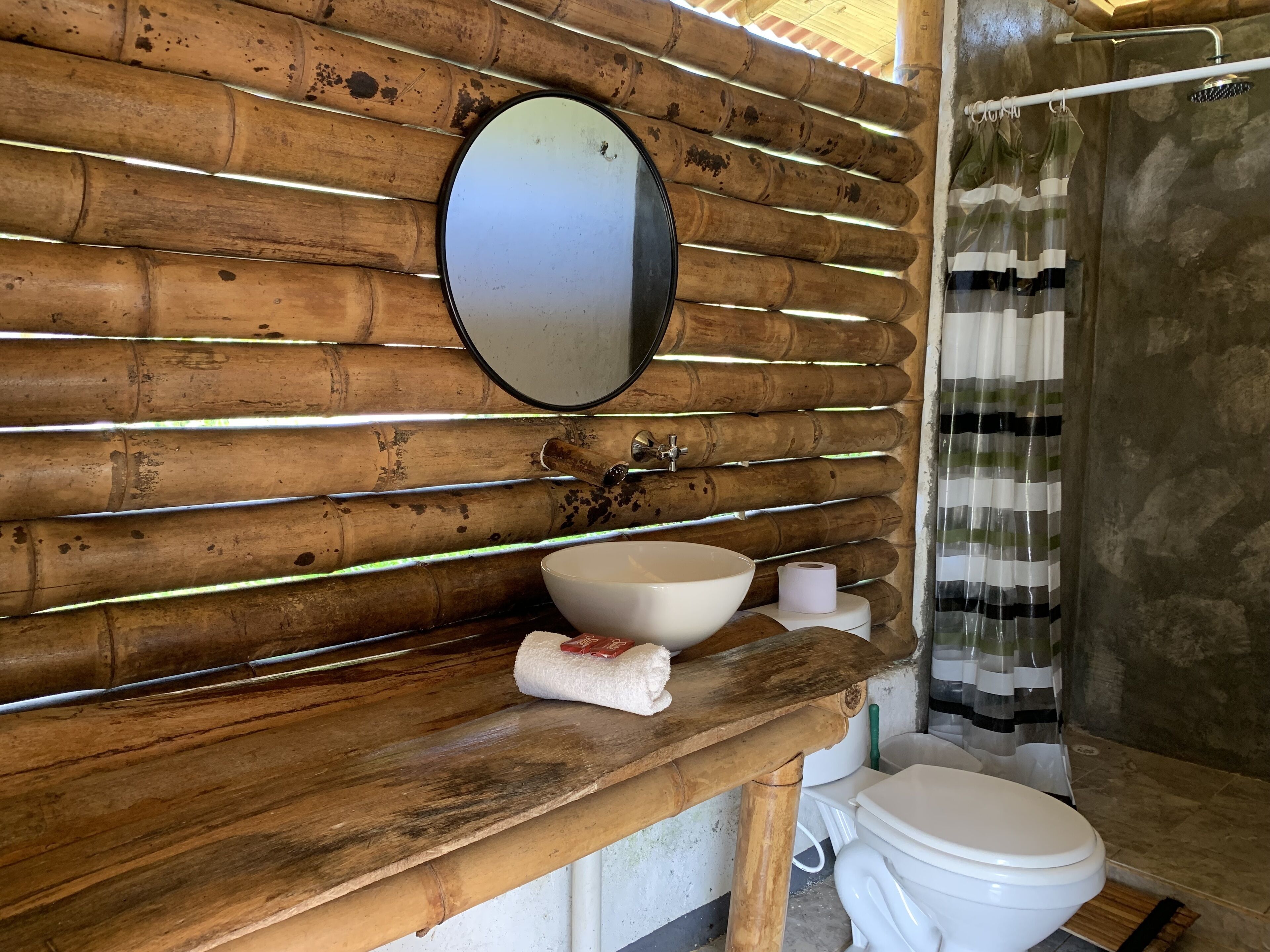 Une salle de bain avec un mur en bois, un miroir rond, un lavabo blanc, des toilettes et un rideau de douche.