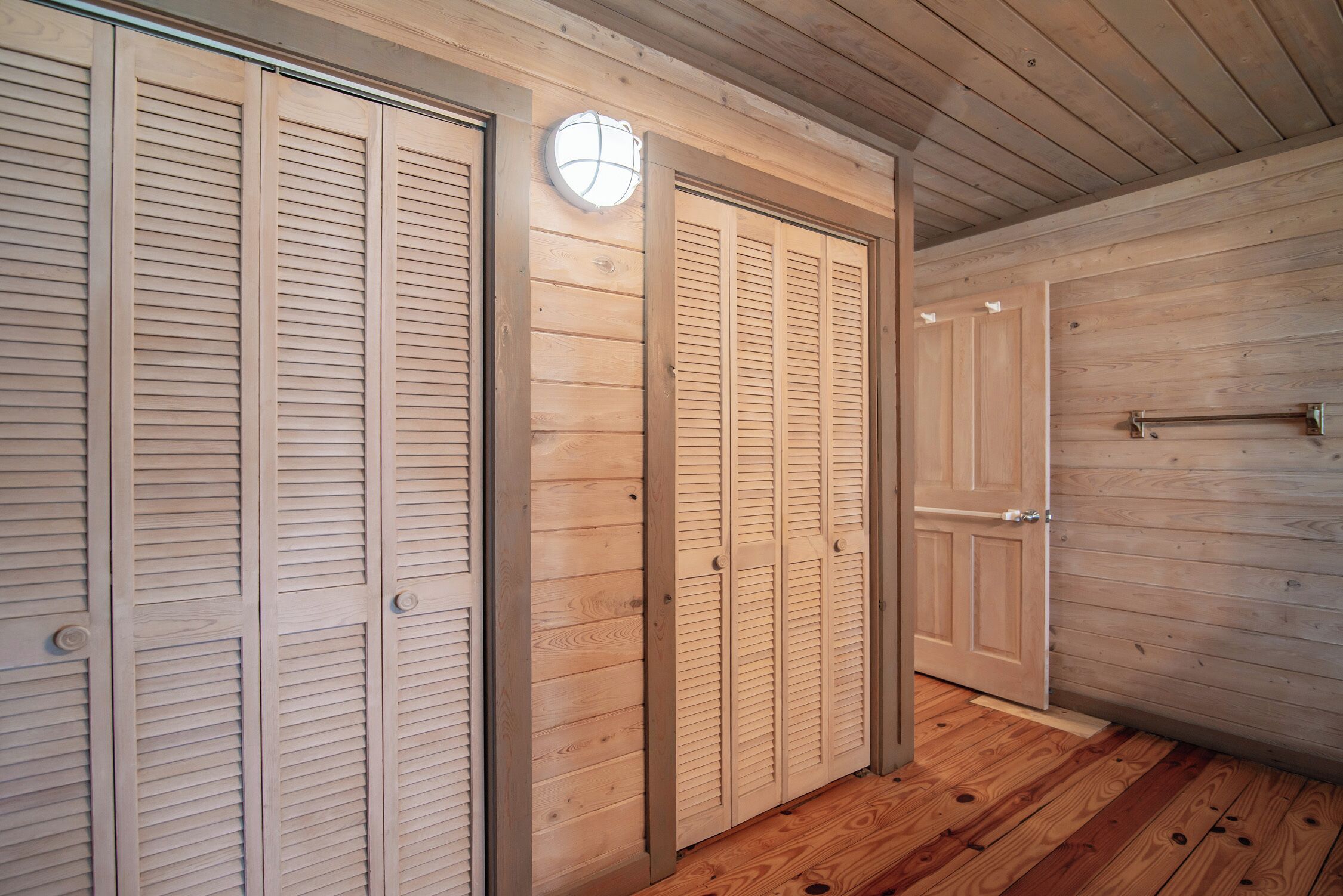 A hallway with wooden panel walls and floors, featuring a set of closed wooden doors with a light fixture above them.