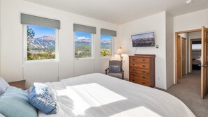 Quarto com uma cama grande, três janelas com vista para as montanhas, cômoda de madeira e uma cadeira.