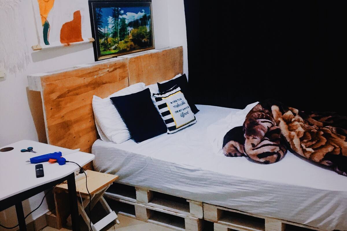 Un dormitorio con estructura de cama de madera, ropa de cama blanca y una manta con estampado de tigre. Hay una mesita con un objeto azul encima, y una estantería de pared con un marco de fotos y un pequeño objeto naranja.