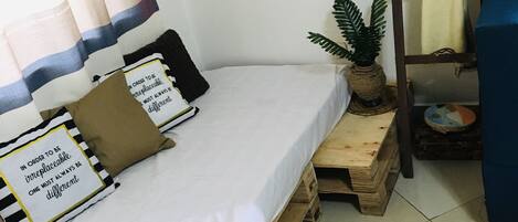 A small, neatly arranged bedroom with a single bed, a small wooden table, a ladder, and a potted plant.