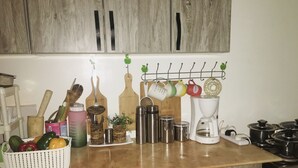 A kitchen with wooden cabinets, a countertop with various kitchen utensils, a coffee maker, and a basket of vegetables. - 250picnics (Kigali)