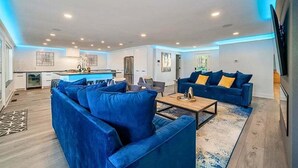 A modern living room with a blue sofa, a coffee table, and a kitchen in the background. - Lakefront Home with Pool, Game Room & Dock (Hot Springs)