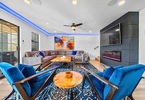 A modern living room with a sectional sofa, blue chairs, a coffee table, a fireplace, and a large TV.