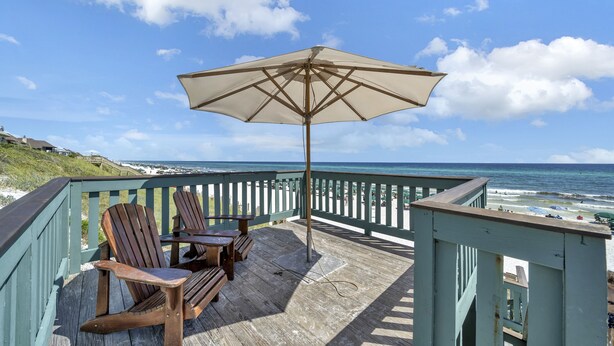 Un pontile in legno con due poltrone Adirondack e un ombrellone grande, con vista su una spiaggia, l'oceano e un cielo sereno.