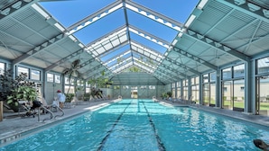 Una piscina interior con techo de cristal, carriles delimitados y sillones de descanso a los lados.
