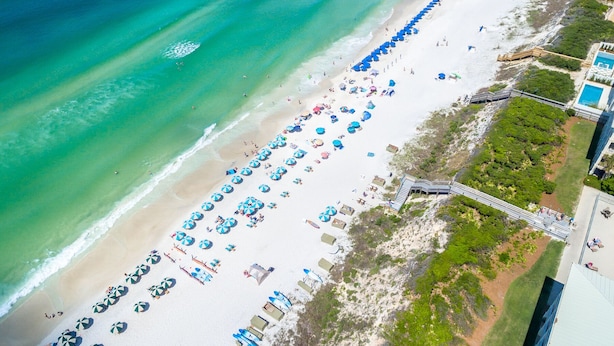 Vista aérea de una playa con hileras de sombrillas y sillas de playa, un paseo de madera y una zona de piscina.