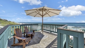 Una terraza de madera con dos sillas Adirondack y un paraguas grande, con vista a una playa, el océano y un cielo despejado.