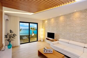 A modern living room with a white sofa, a flat-screen TV, a wooden coffee table, and a view of the ocean through large sliding doors. - ELIA BEACH (Chania)