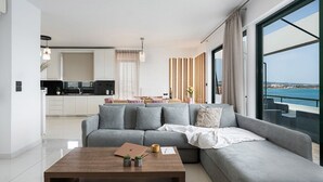 Ein modernes Wohnzimmer mit grauem Sofa, einem Holztisch und einem Laptop. Im Hintergrund ist eine Küche und ein Balkon mit Meerblick zu sehen.