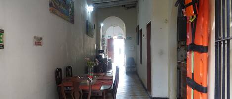 Um corredor com piso xadrez, cadeiras de madeira, uma mesa com um vaso de flores e um quadro na parede.