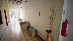 Un pasillo con suelo de cuadros, muebles de mimbre, un jarrón con ramas, un extintor y un letrero de salida de emergencia verde.