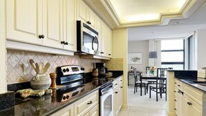 A kitchen with white cabinets, a black countertop, a microwave, and a stove. There is a dining area with a table and chairs in the background. - Beachfront Condo available on a Monthly Basis Only (Marco Island)