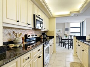 A kitchen with white cabinets, a black countertop, a microwave, and a stove. There is a dining area with a table and chairs in the background. - Beachfront Condo available on a Monthly Basis Only (Marco Island)