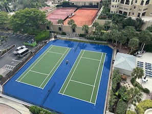 Una cancha de tenis rodeada por un estacionamiento y un edificio con techo rojo.