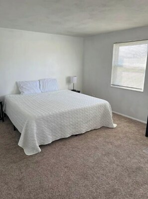 A bedroom with a bed, a nightstand, a lamp, and a window with blinds. - King Beds & Park View, Modern Decor (Vandalia)