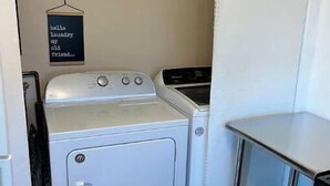 Un lavadero pequeño con lavadora y secadora, un estante con productos de limpieza y un cartel que dice “hola, lavadero, viejo amigo”.
