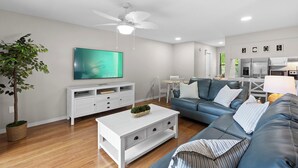Una sala de estar moderna con televisor de pantalla plana, mueble de entretenimiento blanco, sofá azul y ventilador de techo.