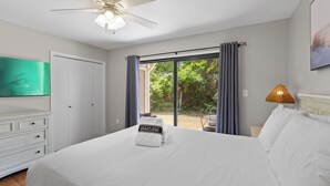 Un dormitorio con cama, ventilador de techo, ventana con cortinas, lámpara de noche y televisor en la pared.