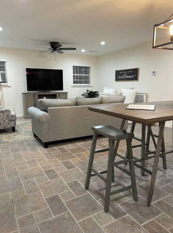 Un salon comprenant un canapé, un téléviseur, un ventilateur de plafond et un coin repas avec une table et des tabourets.