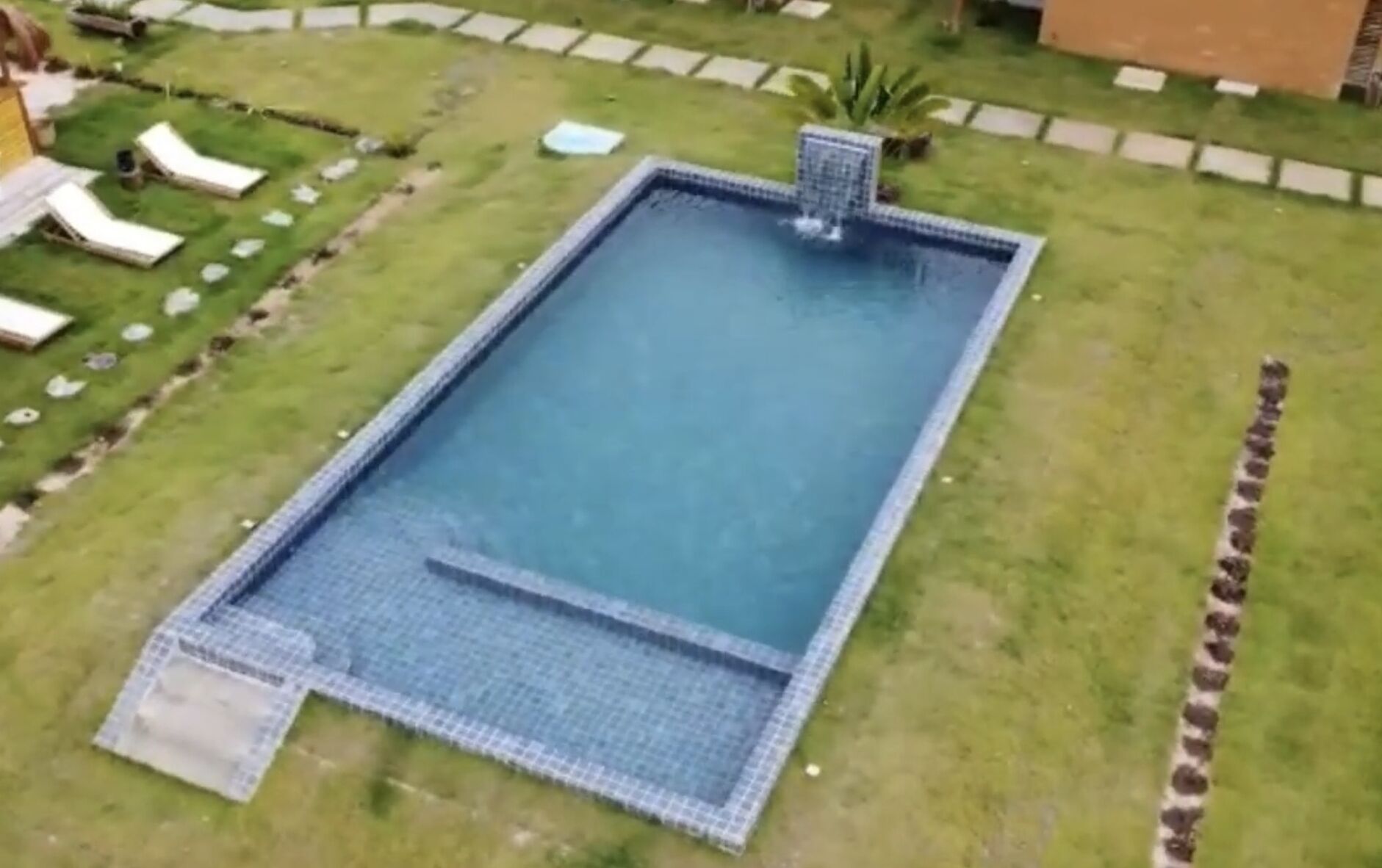 A rectangular swimming pool with a tiled edge, surrounded by a grassy area and a few lounge chairs.