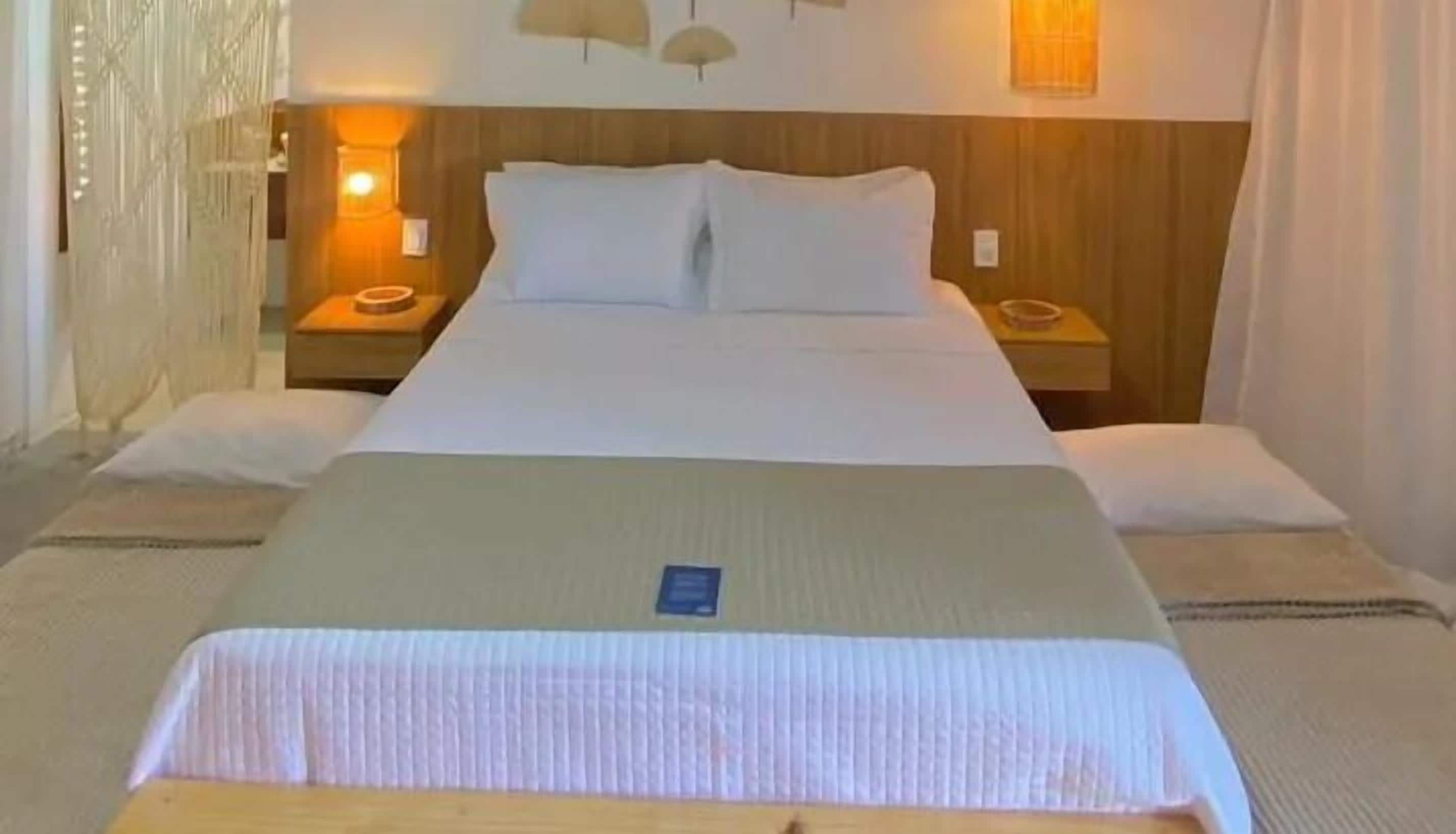 Uma cama bem arrumada com lençóis brancos, duas mesas de cabeceira com abajures e uma cabeceira de madeira.