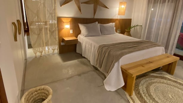 A bedroom with a wooden ceiling, a bed with white bedding, a star decoration, a bedside lamp, and a woven basket. - Pousada Corália (São Miguel dos Milagres)