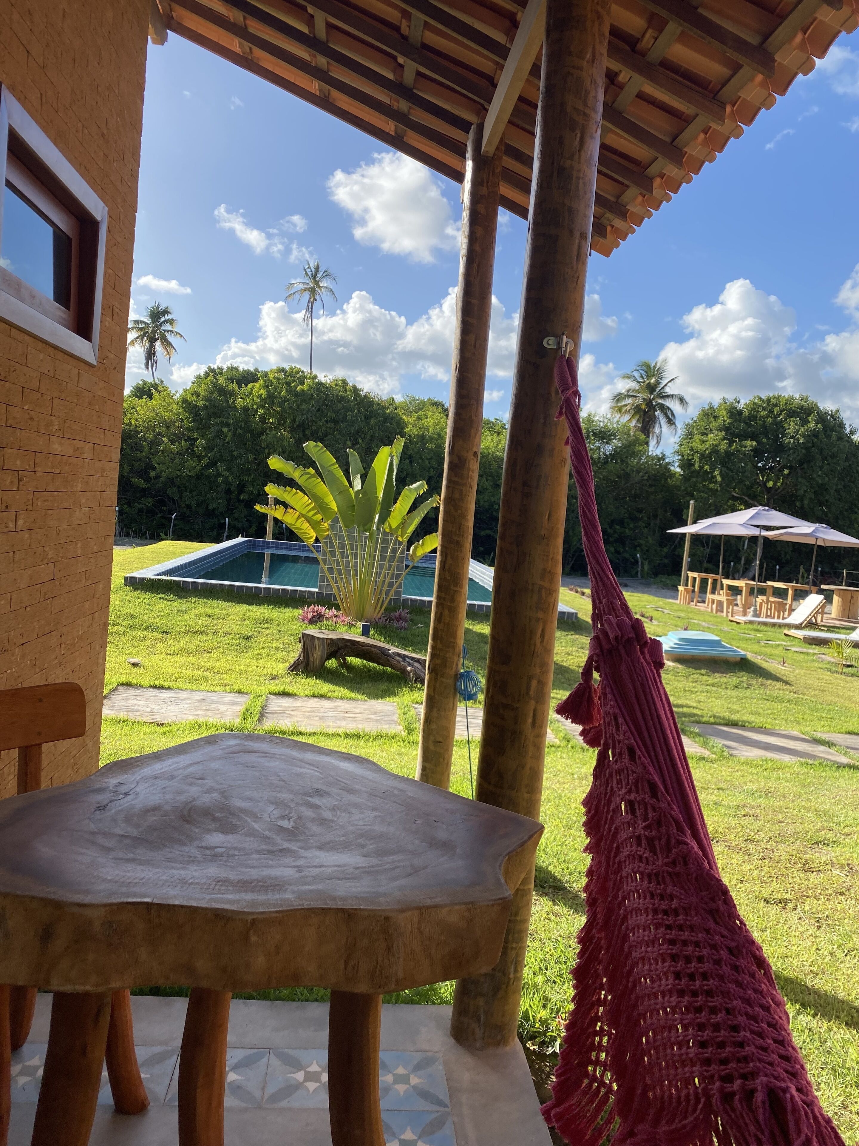 A wooden table under a wooden pergola with a hammock, a pool, and a grassy area with palm trees.