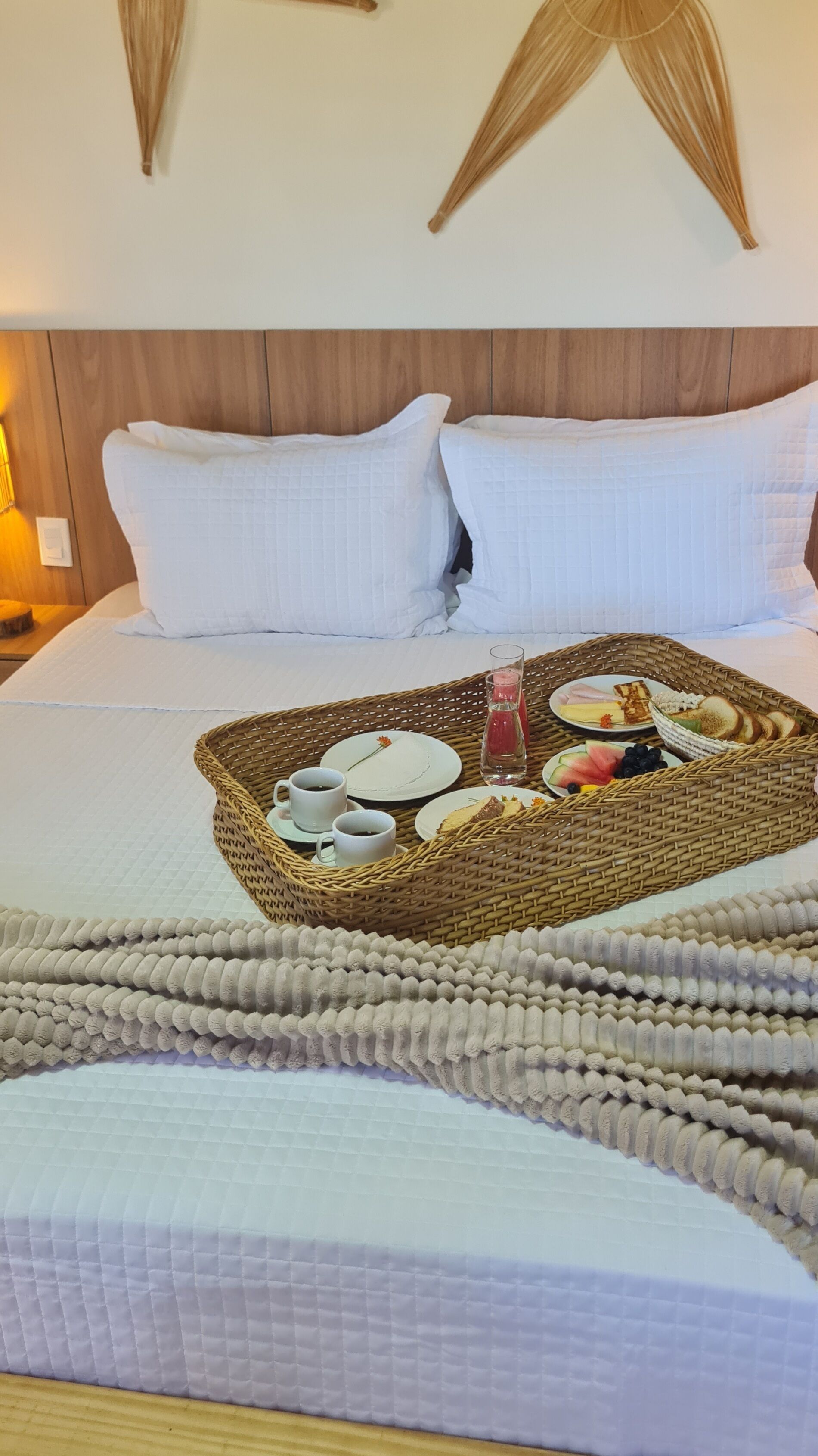 Una cama bien hecha con colcha blanca, dos almohadas blancas y una bandeja tejida con artículos de desayuno como pan, frutas y una botella de agua.