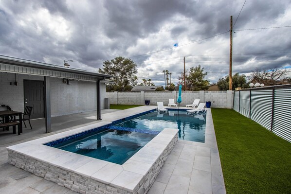 Une piscine rectangulaire dont le bord est recouvert de carreaux, entourée d’un mur en briques blanches et d’une zone herbeuse.