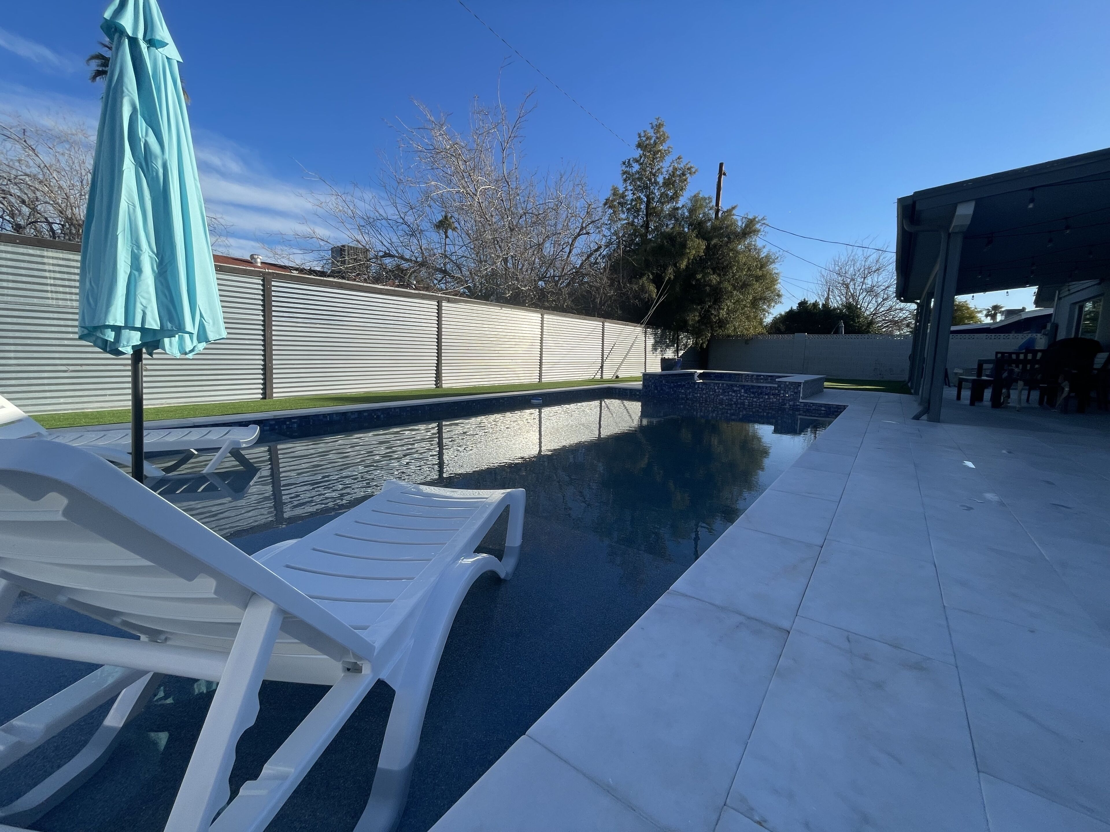 Una piscina con un borde de baldosas, una silla de descanso blanca y una sombrilla azul.
