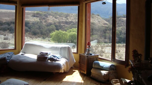 Quarto com cama, uma cadeira e vista para uma paisagem montanhosa através de grandes janelas.