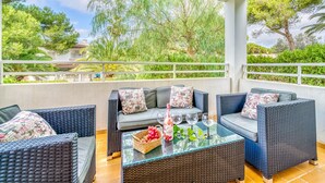 A covered outdoor seating area with wicker furniture and a glass table, overlooking a lush garden and a building in the background. - House with pool Sirenas Playa de Muro (Playa de Muro)