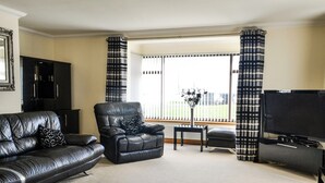 A living room with a black leather sofa, a black leather armchair, a small table with a vase, a flat-screen TV, and a large window with curtains. - Conveniently located just 500 yards from the sandy beach. (Southerness, near Dumfries)