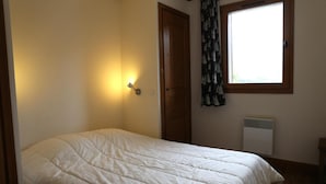 Une chambre à coucher avec un lit, une lampe fixée au mur, une fenêtre avec des rideaux et un radiateur.