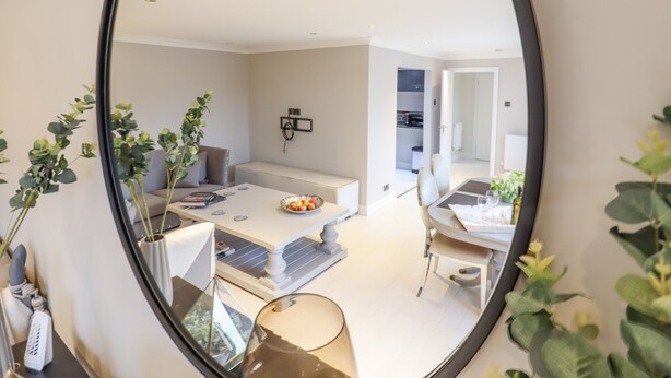 A modern living room with a sofa, coffee table, and a dining area with chairs. There is a kitchen visible in the background. - Closefield (Leighton Buzzard)