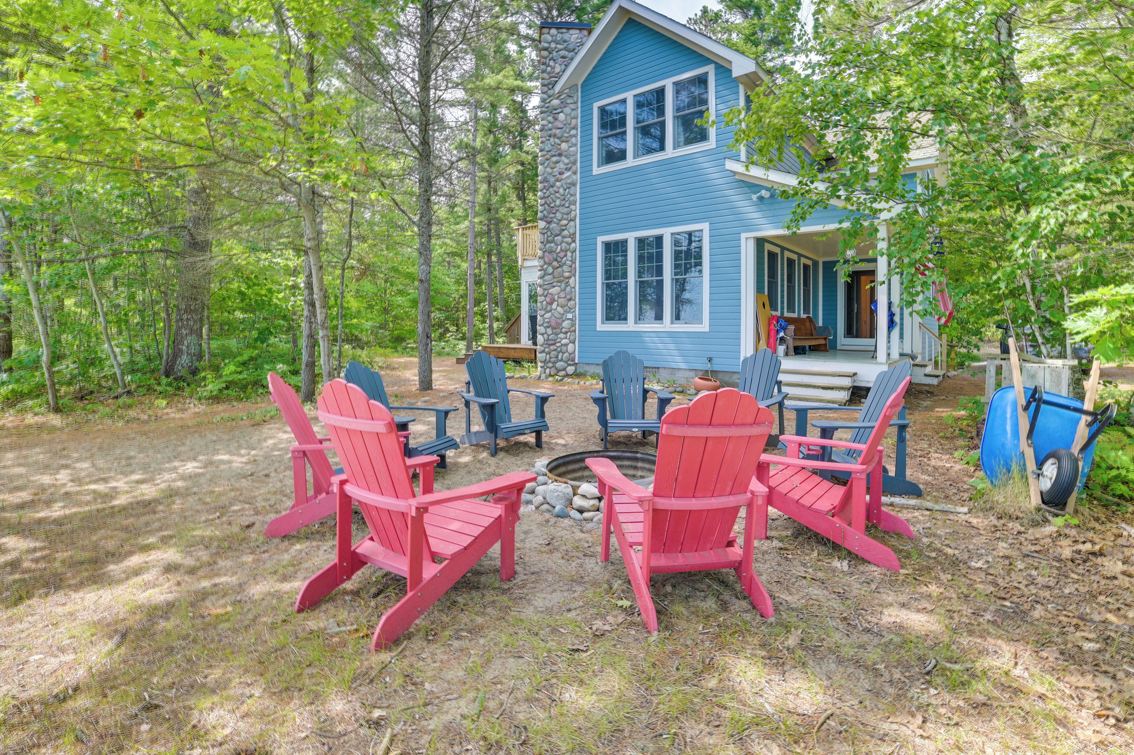 A cabin with a stone exterior, surrounded by a forest. There are red Adirondack chairs and a fire pit in the yard.