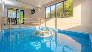 Uma piscina interior com um flutuador inflável colorido, porta de vidro e vista para árvores através de grandes janelas.