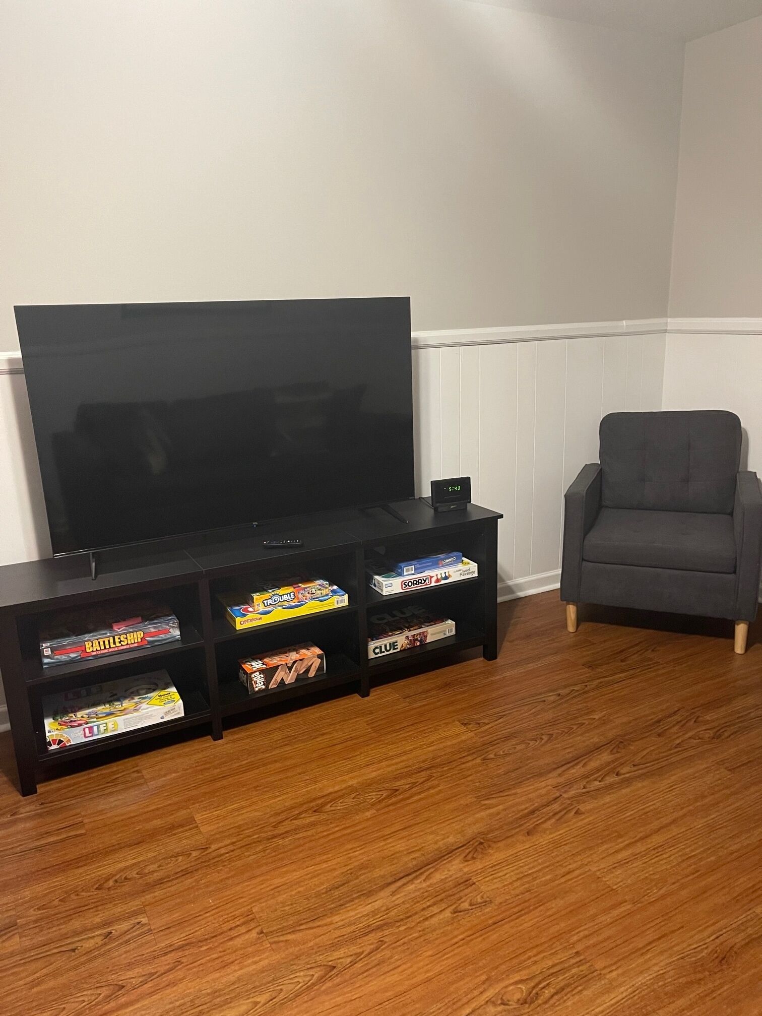 A living room with a flat-screen TV on a black stand, a grey armchair, and a wooden floor.