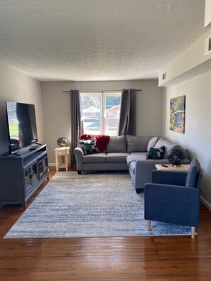 A living room with a grey sofa, a blue armchair, a wooden coffee table, a TV on a cabinet, and a large window with curtains.