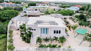 A modern building with solar panels, a thatched-roof structure, and a sandy courtyard with palm trees. - Turibana 50 G (Noord)