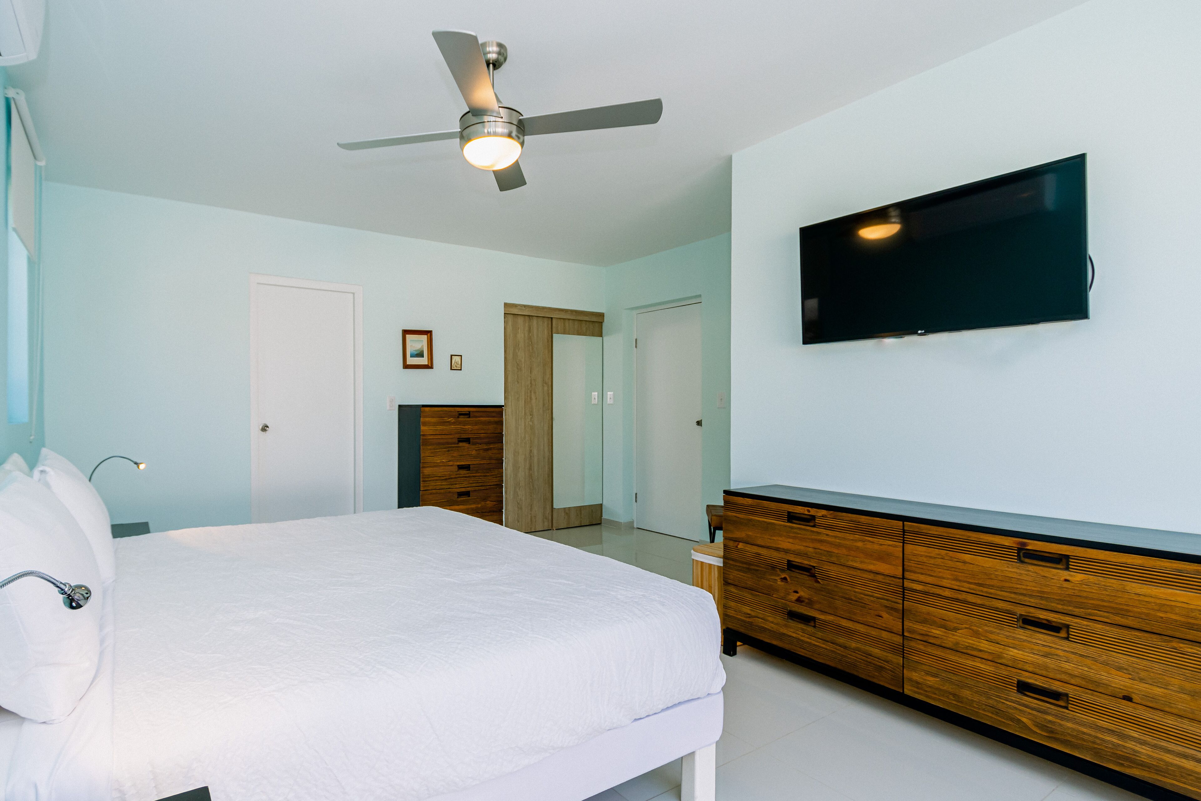 A bedroom with a bed, a wooden dresser, a television mounted on the wall, and a ceiling fan.