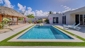 A swimming pool with a thatched roof structure, a patio area with lounge chairs, and a building with a glass door. - Turibana 50 G (Noord)