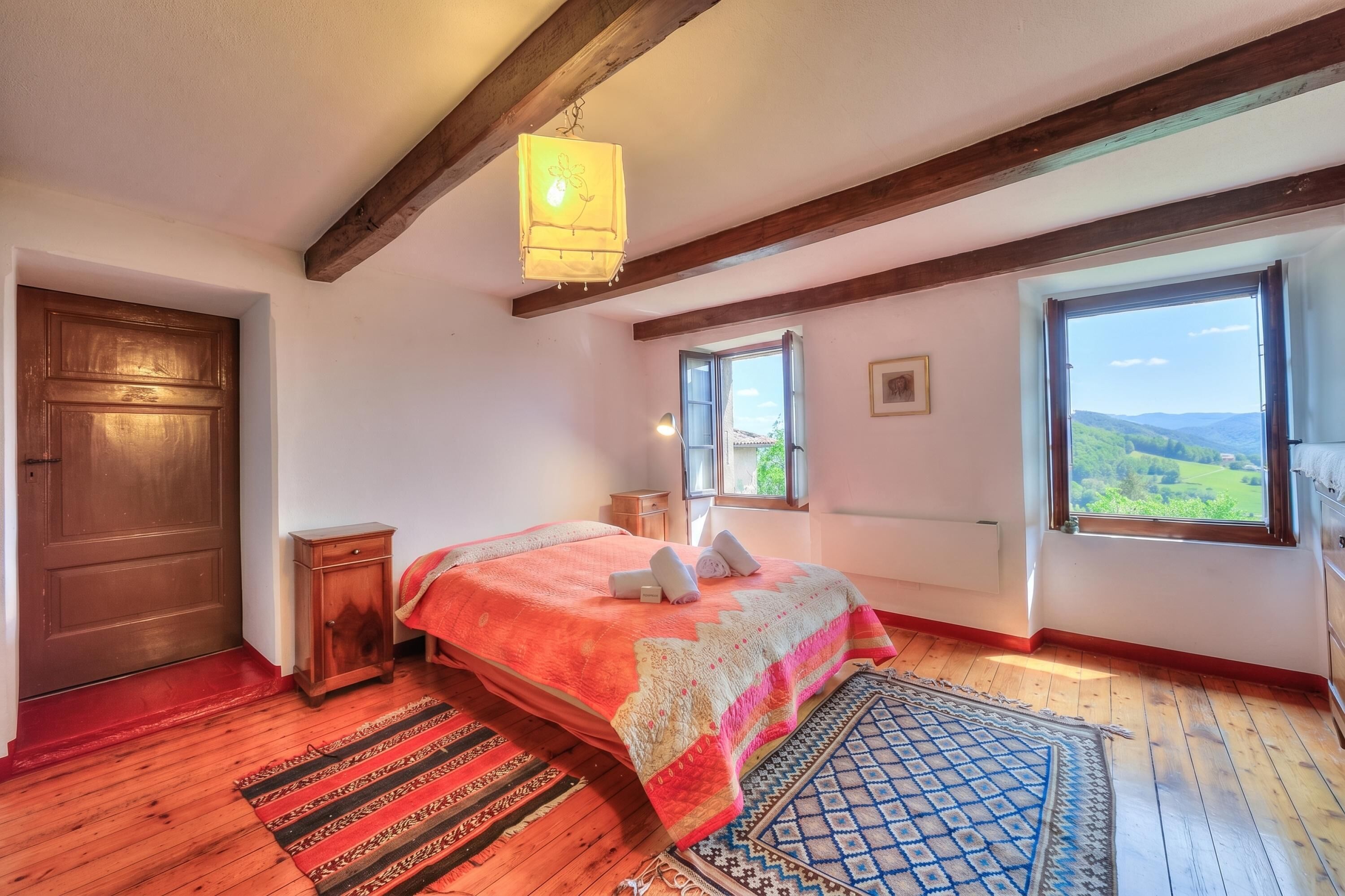 Un dormitorio con piso de madera, cama con manta roja y blanca, cabecera de madera, ventana con vista a follaje, y lámpara colgante.