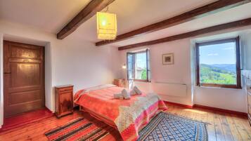 Un dormitorio con piso de madera, cama con manta roja y blanca, cabecera de madera, ventana con vista a follaje, y lámpara colgante.