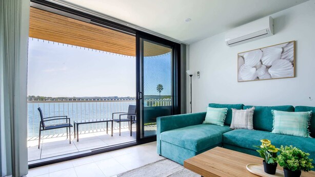 A modern living room with a teal sofa, a wooden coffee table, and a view of the water through large glass doors. - Stunning 1-Bed Bayside Apartment with Superb Views (Batemans Bay)