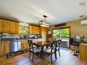 A kitchen with wooden cabinets, a dining table with chairs, a coffee station, and a view through large windows.