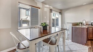 A dining area with a wooden table and white chairs, a kitchen with a wooden countertop, and large windows with curtains.
