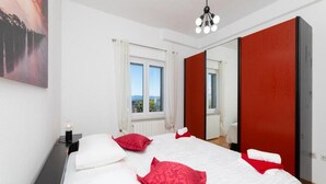 Quarto com cama grande, duas almofadas vermelhas, edredom branco, mesa de cabeceira com abajur, janela com cortinas e guarda-roupa vermelho.