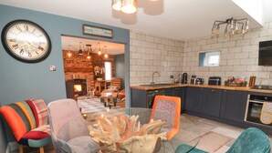 A modern living room with a glass coffee table, colorful chairs, a fireplace, and a kitchen with dark cabinets and a brick wall. - Well Cottage (Beverley)